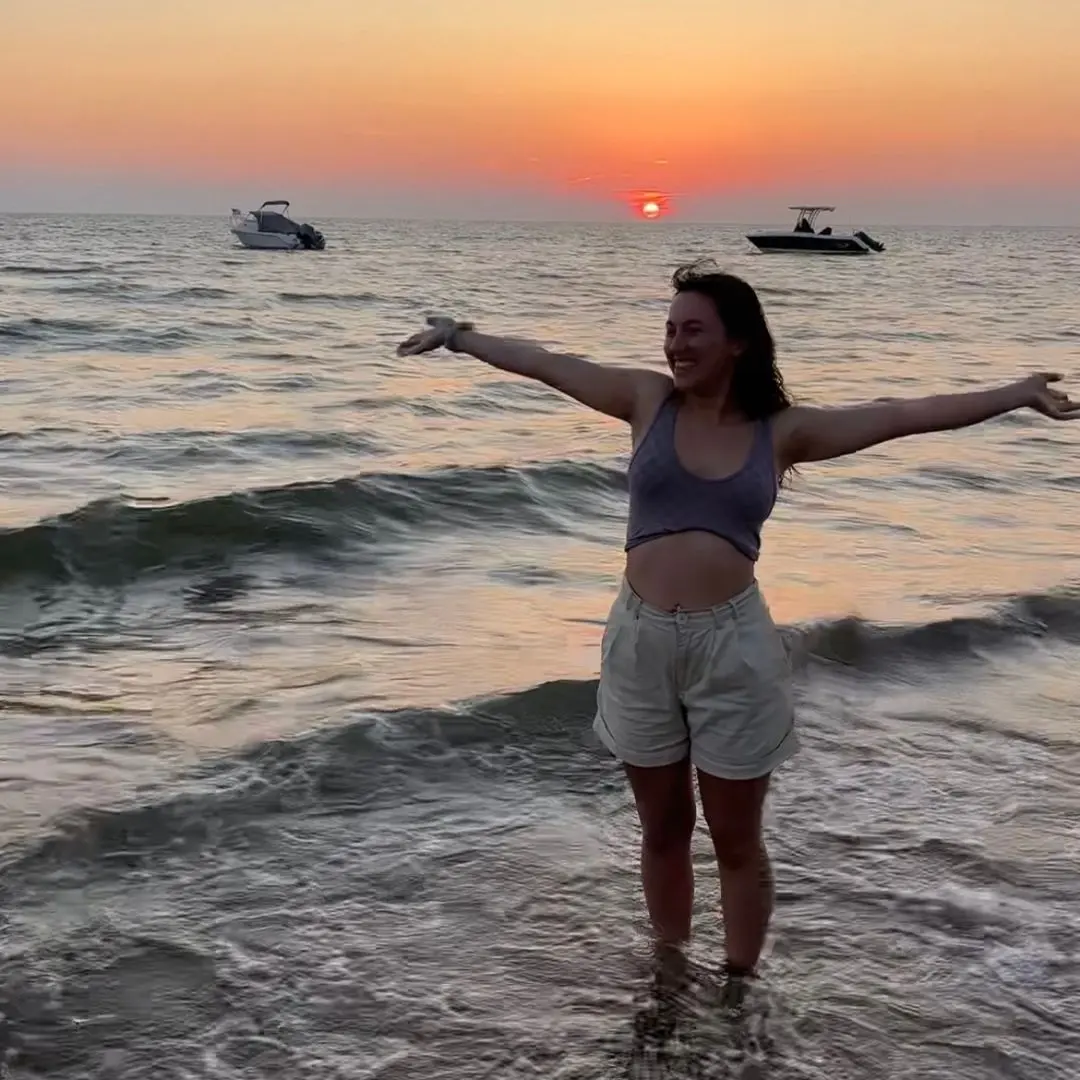 A girl standing in the shallow sea at sunset with her arms out wide, smiling to the camera.