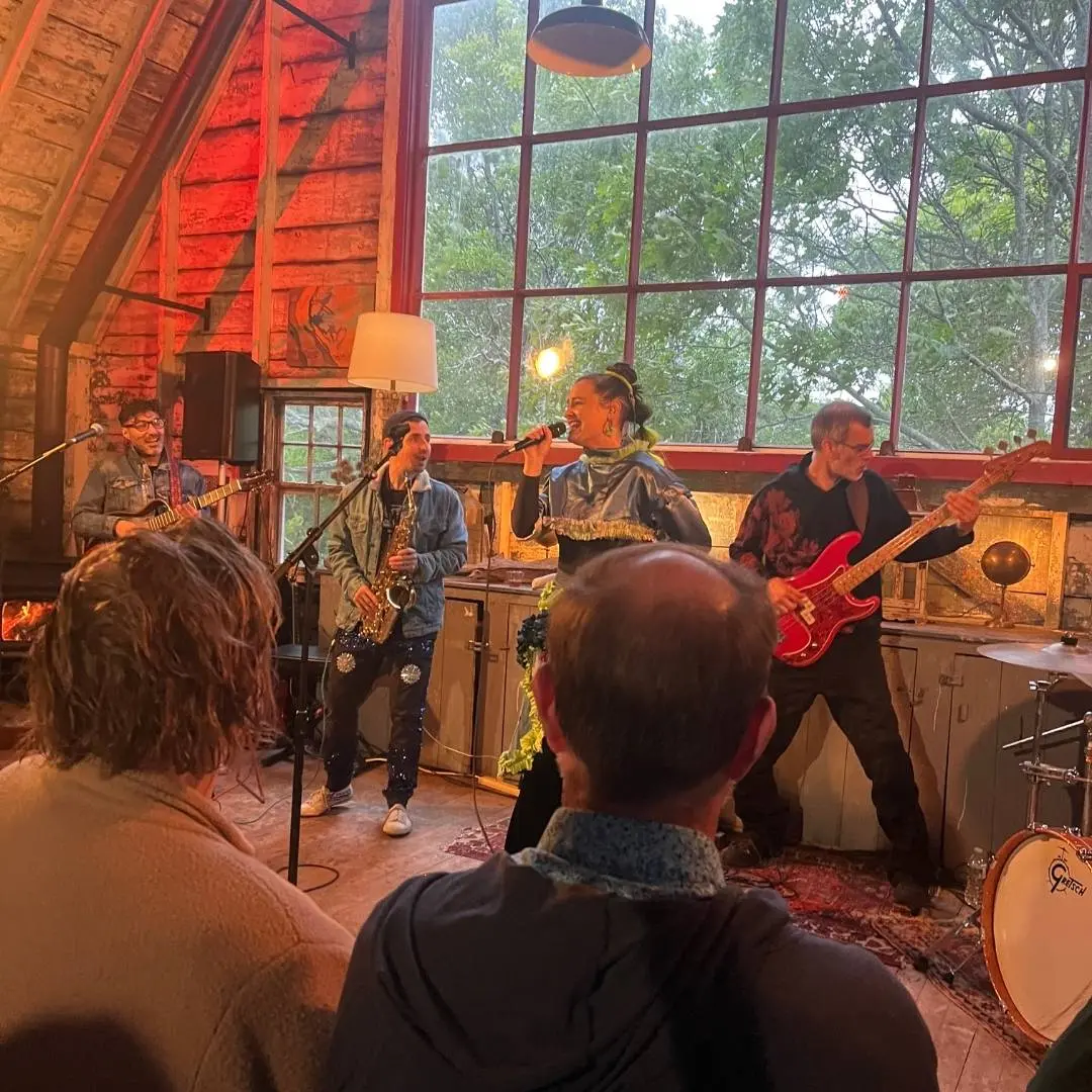 Band playing on a tiny stage in a barn type building in front of a huge window. 