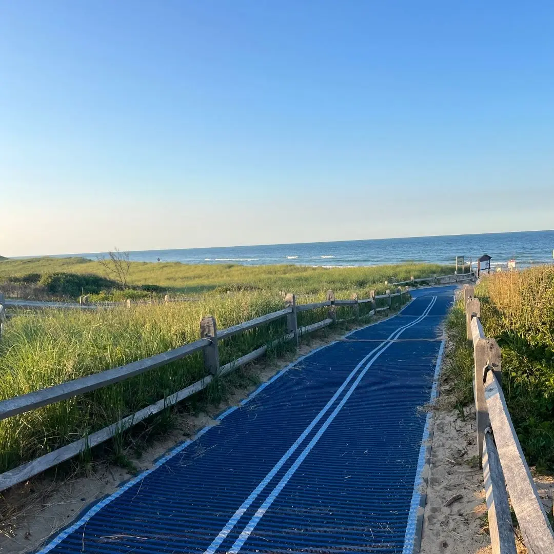 Open tarmac cycle path cut into a field of long grass with the sea ahead. The path leads down to a sandy beach. It's sunset and the sky is clear blue with an orange summery tint.